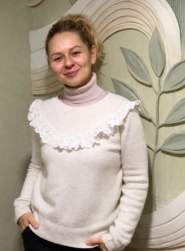 Portrait of artist Daria Romanova standing in front of a sculptural wall relief with botanical forms.