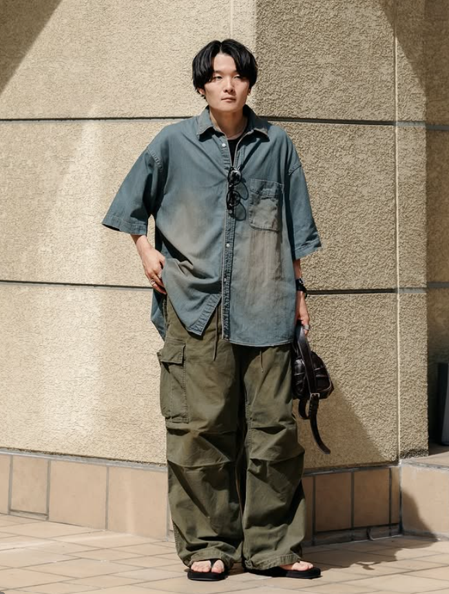 Ryo Takashima wearing an oversized shirt with wide olive cargo trousers, standing against a textured stone wall in an urban setting.