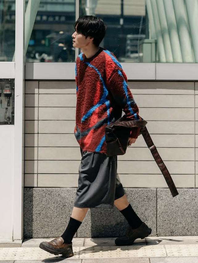 Ryo Takashima walking through an urban space wearing a textured red knit sweater, cropped trousers, and black socks.