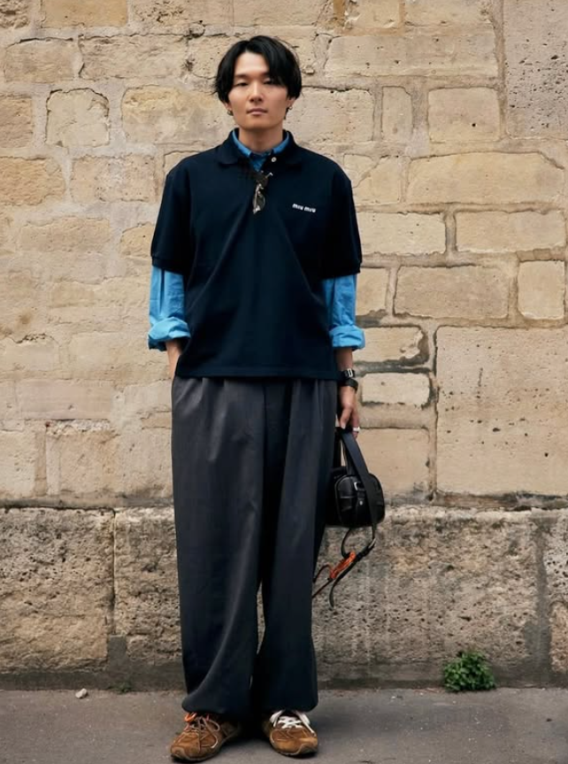 Ryo Takashima wearing a navy top layered over a blue shirt with wide grey trousers, standing against a stone wall.