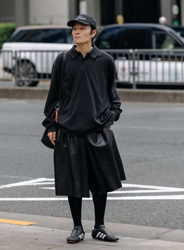Ryo Takashima in a black oversized shirt, layered skirt-style trousers, black cap, and Adidas sneakers, photographed on an urban street.