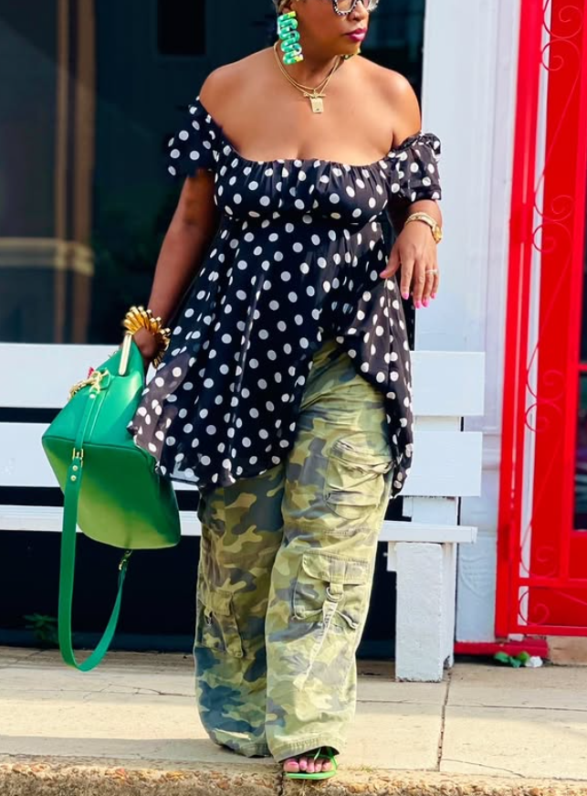 Kenya Freeman wearing a black polka dot off-shoulder top with camouflage trousers, carrying a green handbag in an urban setting.
